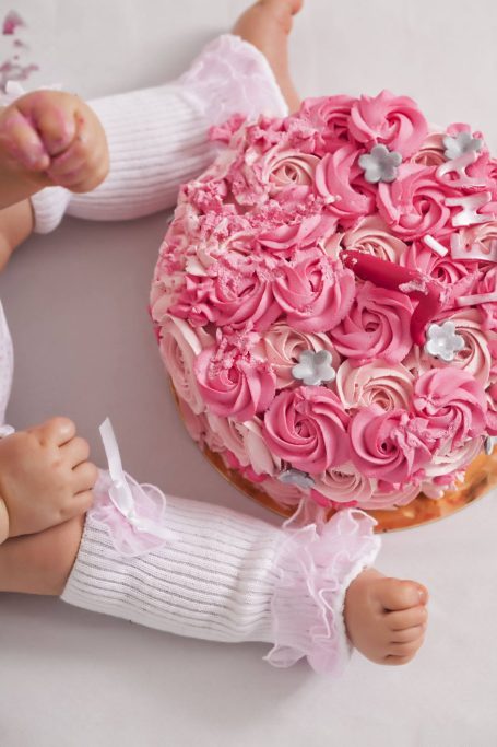 Bébé en chaussettes blanches à côté d'un gâteau rose décoré de crème et de fleurs.